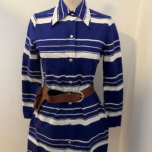 Op de afbeelding: Een vintage blauw-wit gestreepte jurk met een kraag en lange mouwen. De jurk heeft een bruine leren riem en een klein bruin holster. De jurk heeft knopen en twee kleine zakken op de borst.