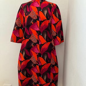 Puede incluir: Un vestido de estilo vintage con cuello alto y mangas tres cuartos. El vestido presenta un vibrante estampado de hojas grandes y estilizadas en tonos de rojo, naranja y burdeos, sobre un fondo negro. Tiene una botonadura frontal.