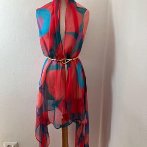 May include: A sheer, colorful scarf with a vibrant pattern of red, blue, and pink circles. The scarf is draped over a mannequin and cinched at the waist with a gold-colored belt. The scarf's design is eye-catching and adds a pop of color.