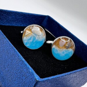 May include: A pair of cufflinks with a beach scene inside. The cufflinks are silver with a clear resin top. The beach scene includes sand, water, and a seashell.