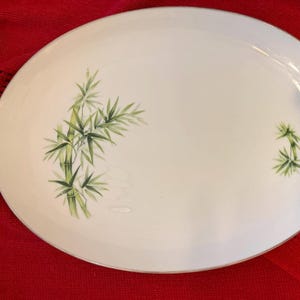 May include: Oval white serving platter with a bamboo design. The platter has a gold rim and green bamboo stalks and leaves painted on the surface. The platter is on a red surface.