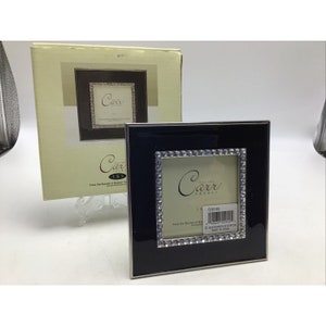 Puede incluir: Un marco de fotos cuadrado negro con un borde plateado y una fila de pequeños cristales transparentes alrededor del borde interior. El marco está junto a su caja original, que también presenta el diseño del marco. El marco mide 7,6 x 7,6 cm.