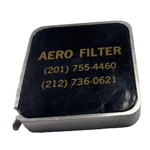 Puede incluir: Una cinta métrica de metal negro con el texto "AERO FILTER (201) 755-4460 (212) 736-0621" impreso en dorado.