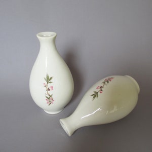 May include: Two white ceramic vases with a teardrop shape. Each vase features a delicate floral design with pink blossoms and green leaves. One vase is upright, the other is lying on its side.