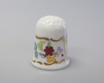May include: A white porcelain thimble with a floral design. The thimble features a gold trim and colorful flowers in red, yellow, blue, and green. The thimble is a sewing accessory.