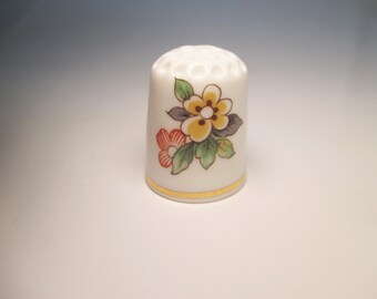 May include: White ceramic thimble with a floral design. The thimble features a yellow and red flower with green leaves. A gold band encircles the base. The top has small holes for ventilation. A decorative sewing accessory.