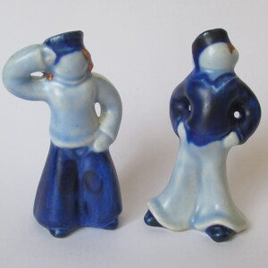 May include: Two ceramic figurines of people in sailor-style outfits. One figure is light blue with a dark blue skirt, and the other is dark blue with a light blue skirt. Both have dark blue hats and are in a pose.