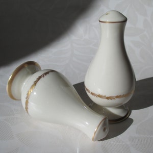 May include: Two white ceramic salt and pepper shakers with gold trim and a gold leaf design. One shaker is upright, the other is lying on its side. The shakers have a classic, elegant design.