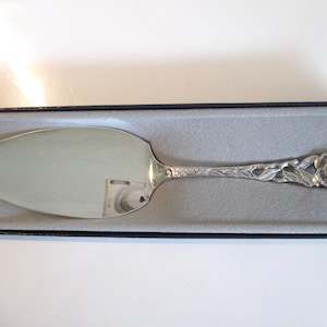 May include: Silver-colored cake server with a floral design on the handle. The server is in a black box with a gray interior. The handle features a rose and leaf pattern, and the blade is smooth and reflective.