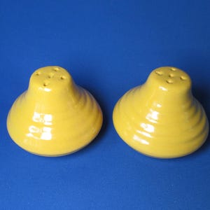 May include: Two yellow ceramic salt and pepper shakers. Each shaker has a bulbous base with ridged detailing and a top with four holes. The shakers are set against a solid blue background.
