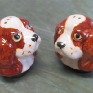 May include: A pair of ceramic salt and pepper shakers shaped like dog heads. The shakers are white with brown markings and black noses. Each shaker has three holes on top for dispensing salt or pepper.
