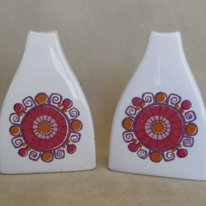May include: Two white ceramic vases with a geometric design in shades of pink, orange, and purple. The design features a circular pattern with swirls and dots.