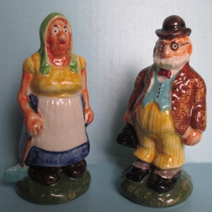 May include: A pair of ceramic figurines depicting a woman and a man. The woman is wearing a green and yellow dress with a white apron and holding a shovel. The man is wearing a brown jacket, yellow pants, and a black hat. Both figurines have painted details and stand on green bases.