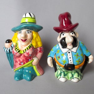 May include: A pair of whimsical ceramic figurines. One is a woman with a yellow wig, a turquoise hat, and a red floral dress. The other is a man with a burgundy hat, a blue shirt, and a black mustache. Both have exaggerated features.