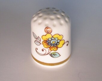 May include: White ceramic thimble with a yellow and blue floral design. The thimble has a gold band around the base and small holes at the top. A decorative sewing accessory.