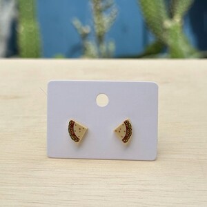 May include: A pair of small stud earrings shaped like tacos, featuring a beige shell, green lettuce, and red tomato details. The earrings are attached to a rectangular white display card with a circular hole at the top for hanging purposes.