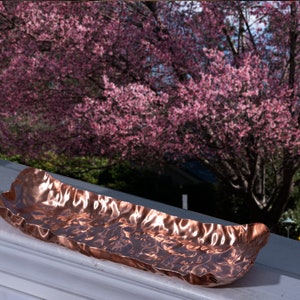 Puede incluir: Un plato rectangular de color cobre con una superficie texturizada y ondulada. El plato est&aacute; colocado en un alf&eacute;izar blanco, con un vibrante &aacute;rbol de flores rosas visible a trav&eacute;s de la ventana en el fondo.