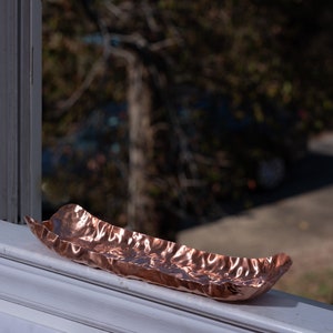 Puede incluir: Una bandeja decorativa oblonga de color cobre con una superficie texturizada y ondulada. La bandeja est&aacute; colocada en un alf&eacute;izar blanco, con una escena exterior borrosa visible en el fondo. La bandeja mide unos 30 cm de largo.