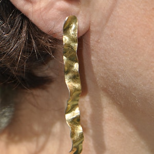 May include: A pair of long, gold earrings with a wavy, textured design. The earrings are hanging from a person's earlobe.