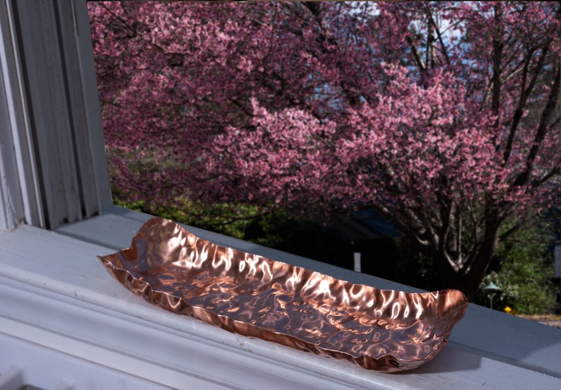 Puede incluir: Un plato rectangular de color cobre con una superficie texturizada y ondulada. El plato est&aacute; colocado en un alf&eacute;izar blanco, con un vibrante &aacute;rbol de flores rosas visible a trav&eacute;s de la ventana en el fondo.