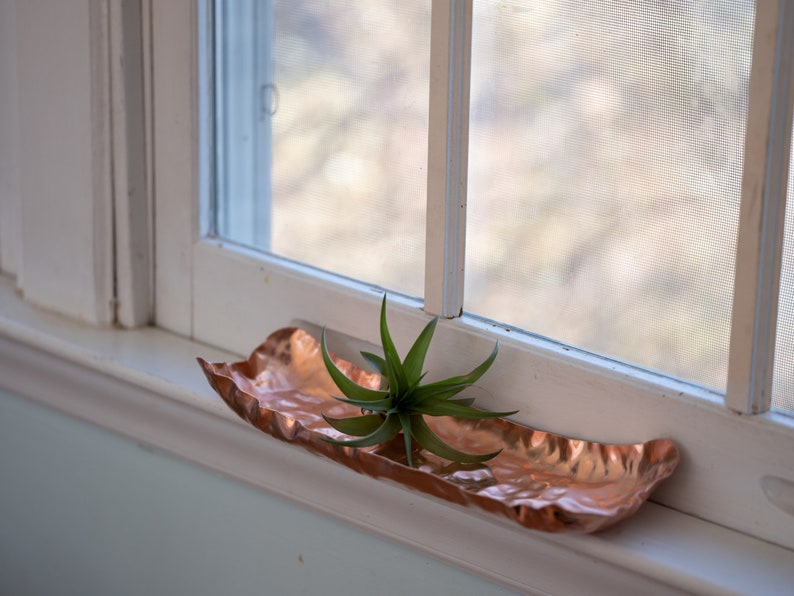 Puede incluir: Una bandeja decorativa oblonga de color cobre con una superficie texturizada. La bandeja contiene una peque&ntilde;a planta a&eacute;rea verde. La bandeja est&aacute; colocada en un alf&eacute;izar blanco, con una ventana al fondo.