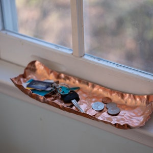 Puede incluir: Una bandeja de cobre martillado con borde ondulado sobre un alf&eacute;izar. La bandeja contiene llaves, tarjetas y monedas. El cobre tiene un color marr&oacute;n rojizo c&aacute;lido y una superficie texturizada. La bandeja mide unos 30 cm de largo.