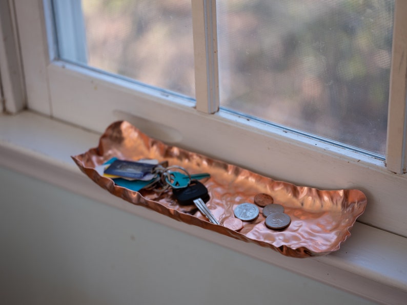 Puede incluir: Una bandeja de cobre martillado con borde ondulado sobre un alf&eacute;izar. La bandeja contiene llaves, tarjetas y monedas. El cobre tiene un color marr&oacute;n rojizo c&aacute;lido y una superficie texturizada. La bandeja mide unos 30 cm de largo.