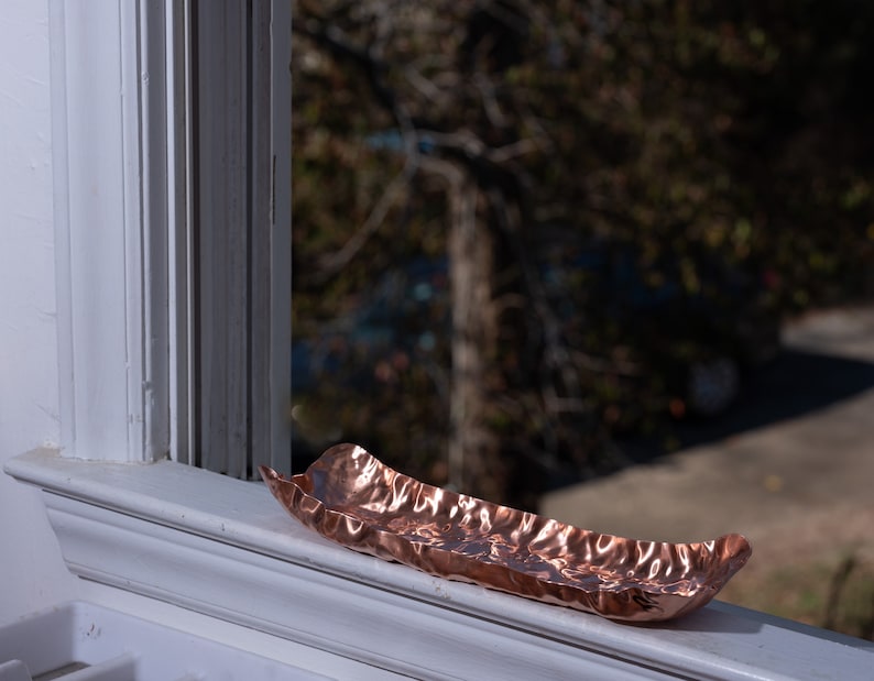 Puede incluir: Una bandeja decorativa oblonga de color cobre con una superficie texturizada y ondulada. La bandeja est&aacute; colocada en un alf&eacute;izar blanco, con una escena exterior borrosa visible en el fondo. La bandeja mide unos 30 cm de largo.