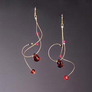 May include: Elegant gold-tone wire earrings with a flowing design. Each earring features a large, teardrop-shaped, dark red crystal, accented by smaller red, pink, and black beads. The earrings have a hook closure.