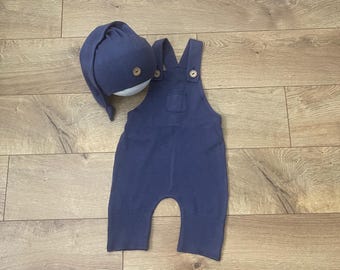 Puede incluir: Conjunto de ropa de bebé azul marino, que incluye un gorro de punto y un peto. El gorro y el peto tienen botones de madera. El peto tiene un pequeño bolsillo en el pecho. El conjunto está hecho de un tejido de punto suave.