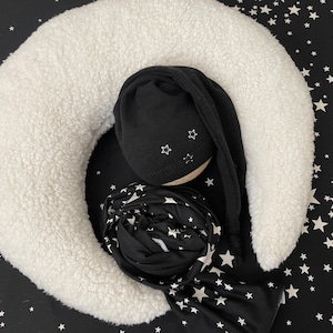 Pode incluir: Uma almofada branca em forma de lua crescente com um tecido estampado de estrelas pretas e brancas e um chapéu de malha preto com estrelas brancas.