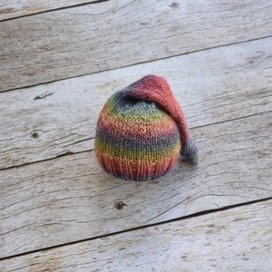 May include: A colorful knitted baby hat with a long, pointed top. The hat is striped in shades of red, orange, yellow, green, blue, and purple.