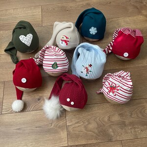 May include: A collection of whimsical Christmas gnome hats in various colors and designs. The hats include red, green, and blue, with festive embellishments like hearts, snowflakes, and snowmen. Some hats have long, pointed tops, while others feature pom-poms.