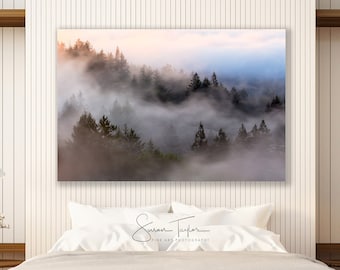 Puede incluir: Una cama blanca con un edredón beige y almohadas blancas en un dormitorio moderno con una gran fotografía enmarcada de una escena de bosque brumoso sobre la cabecera. El dormitorio tiene paredes blancas, suelos de madera y dos mesitas de noche con lámparas blancas y flores.