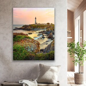 May include: A white lighthouse stands tall on a rocky cliff overlooking a calm ocean at sunset. The sky is a soft pink and orange, and the water is a deep blue. The lighthouse is surrounded by green vegetation and rocks.