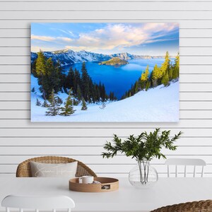 May include: A panoramic view of Crater Lake in Oregon, with snow-covered mountains and a blue lake in the background. The lake is surrounded by evergreen trees and a small island in the center. The sky is a bright blue with white clouds.