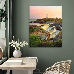 May include: A white lighthouse stands on a rocky cliff overlooking a calm ocean at sunset. The sky is a soft pink and orange, and the water is a deep blue. The lighthouse is surrounded by green grass and rocks.