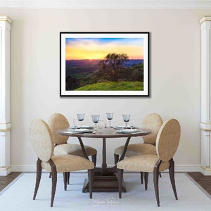 May include: A framed photograph of a landscape scene with a lone tree in the foreground. The sky is a vibrant mix of orange, pink, and purple hues, with a golden glow emanating from the horizon. The hills in the background are a soft purple and green.