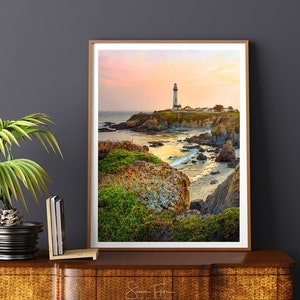 May include: A white lighthouse stands on a rocky cliff overlooking a calm ocean at sunset. The sky is a soft pink and orange, and the water is a deep blue. The lighthouse is surrounded by green grass and rocks.