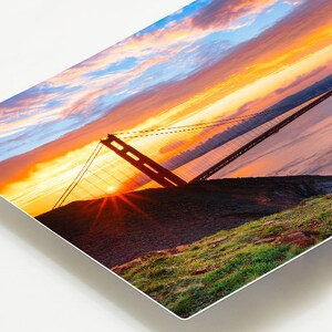 K&ouml;nnte beinhalten: Ein Foto der Golden Gate Bridge in San Francisco, Kalifornien. Die Br&uuml;cke ist vor einem leuchtend orangefarbenen und rosafarbenen Sonnenuntergangshimmel silhouettiert. Die Sonne geht hinter der Br&uuml;cke unter und wirft einen warmen Schein auf die Szene.