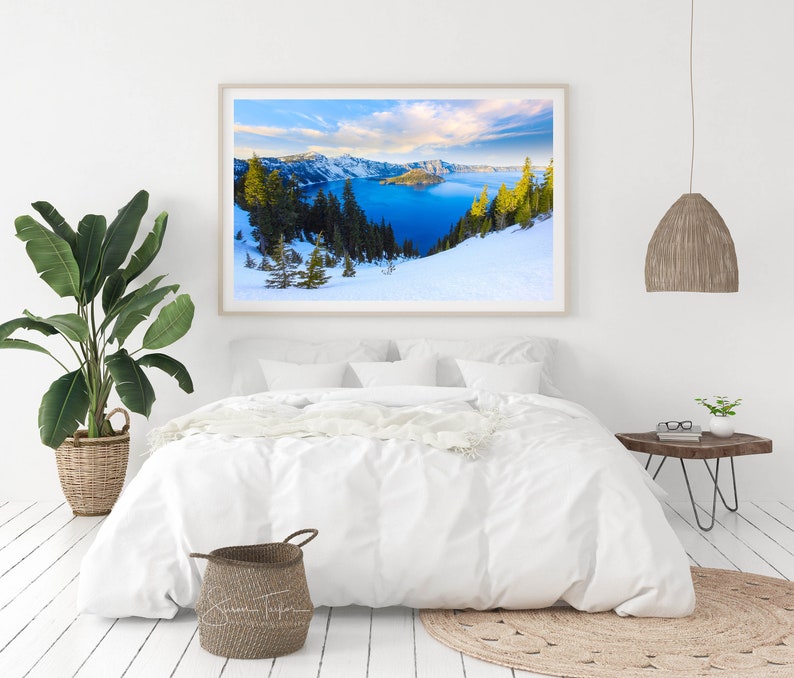 May include: A framed photograph of a scenic landscape with a lake surrounded by snow-capped mountains and evergreen trees. The lake is a vibrant blue, and the sky is a clear blue with white clouds. The photo is displayed on a white wall above a white bed with a white duvet and a white throw blanket.