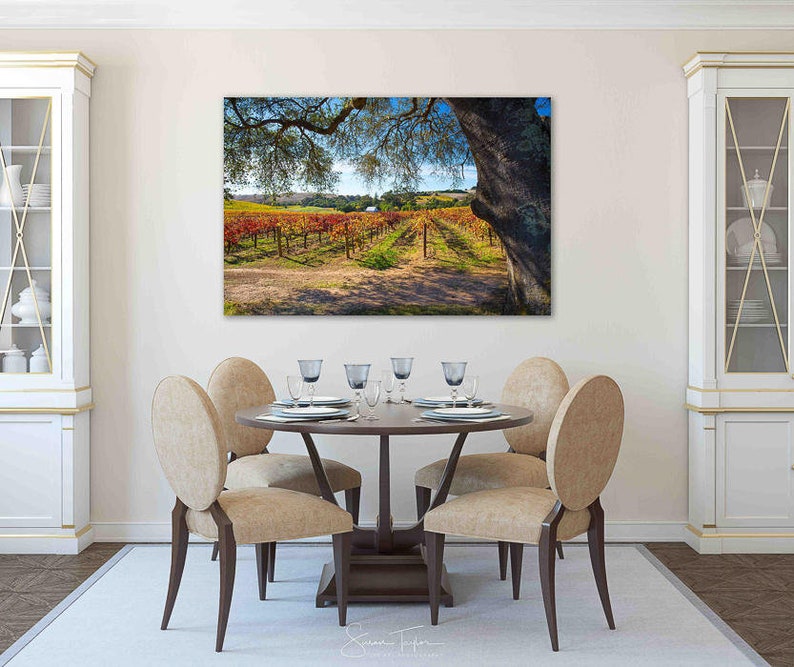 May include: A dining room with a wooden table and six chairs. The table is set with plates, silverware, and glasses. A large framed photograph of a vineyard with a tree in the foreground hangs on the wall behind the table.