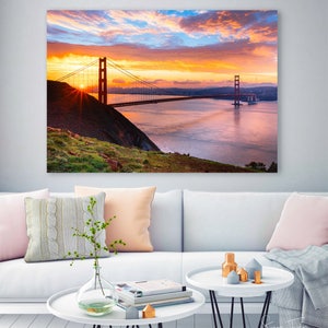 K&ouml;nnte beinhalten: Ein Panoramablick auf die Golden Gate Bridge in San Francisco, Kalifornien. Die Br&uuml;cke ist vor einem leuchtend orangefarbenen und rosafarbenen Sonnenuntergangshimmel mit fluffigen Wolken silhouettiert. Die Sonne geht hinter der Br&uuml;cke unter und wirft einen warmen Schein auf das Wasser.