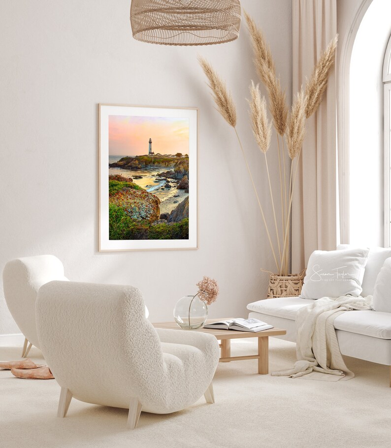 May include: A framed photograph of a lighthouse on a rocky coastline with a sunset sky in the background. The photo is hanging on a white wall in a living room with white furniture and a vase of flowers on a coffee table.