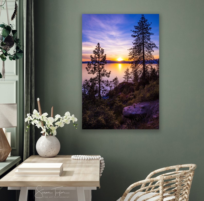 May include: A scenic photograph of a lake at sunset with two silhouetted trees in the foreground. The sky is a vibrant mix of purple, pink, and orange hues. The water reflects the colors of the sky, creating a serene and tranquil scene.