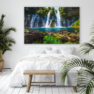 May include: A white bed with a white blanket and a wooden bench in front of it. A large photograph of a waterfall cascading down a rocky cliff with lush green foliage is hanging on the wall behind the bed.