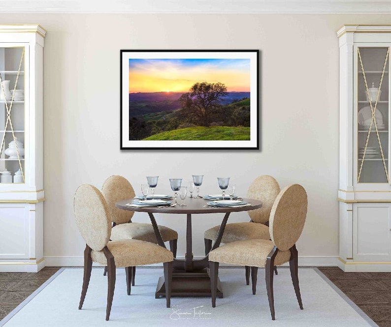 May include: A framed photograph of a landscape scene with a lone tree in the foreground. The sky is a vibrant mix of orange, pink, and purple hues, with a golden glow emanating from the horizon. The hills in the background are a soft purple and green.