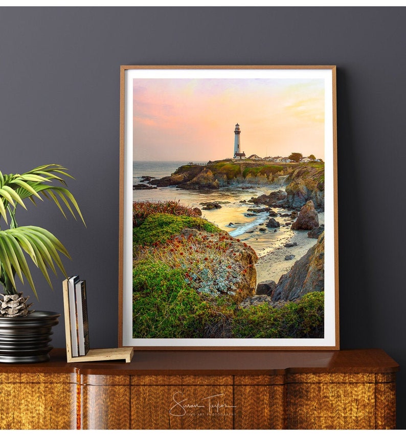 May include: A white lighthouse stands on a rocky cliff overlooking a calm ocean at sunset. The sky is a soft pink and orange, and the water is a deep blue. The lighthouse is surrounded by green grass and rocks.
