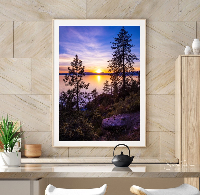 May include: A framed photograph of a sunset over a lake with two silhouetted trees in the foreground. The sky is a vibrant mix of purple, pink, and orange.