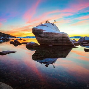 Puede incluir: Una gran formaci&oacute;n rocosa con un peque&ntilde;o &aacute;rbol creciendo en la cima, cubierta de nieve, se encuentra en un lago tranquilo al atardecer. El agua refleja el cielo colorido, creando una hermosa escena.